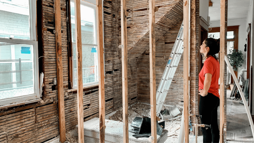 A woman inspecting the exposed wall studs and original lath framing of a historic home during a renovation, assessing the structural integrity before remodeling.